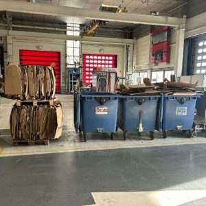 Intérieur d'un atelier industriel avec à gauche une balle de cartons compactés sur palette et à droite trois bennes de collecte bleues remplies de déchets carton en vrac — illustration du contraste avant/après compactage.