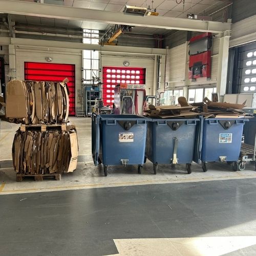 Intérieur d'un atelier industriel avec à gauche une balle de cartons compactés sur palette et à droite trois bennes de collecte bleues remplies de déchets carton en vrac — illustration du contraste avant/après compactage.